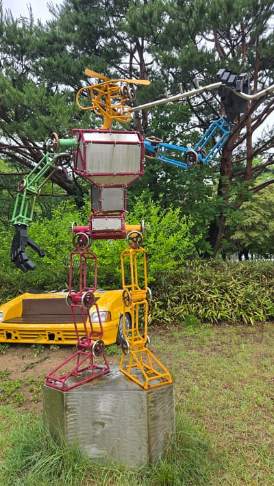 An outdoor metal sculpture resembling a robot is situated in a park in Gwangmyeong, South Korea. The robot is constructed from various colored metal components, including a yellow head with propellers, a red and silver torso, green and blue arms, and red and yellow legs. The robot stands on a metallic cube base, which is surrounded by grass and vegetation. In the background, a yellow car is partially visible, along with trees and bushes. The sky is overcast, suggesting a cloudy day. There are no people or animals present in the image.