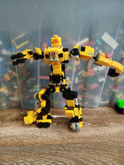 A yellow and black Lego robot is standing on a wooden surface. The robot has a blocky build with arms, legs, and a torso. Its arms are extended outwards, and its legs are positioned as if it's about to move. The robot is predominantly yellow with black accents, including wheels on its feet and hands. The background is a transparent plastic bin filled with various Lego bricks in different colors and sizes. The lighting appears to be indoor, suggesting the image was taken during the day or under artificial light. No text is visible in the image. The setting appears to be a personal space, possibly a playroom or a hobby area.