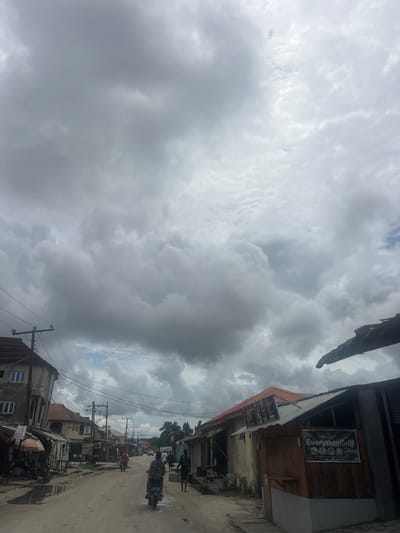 The image depicts a street scene in Awoyaya, Nigeria, under a cloudy sky. The unpaved road shows puddles of water, suggesting recent rain. On the left side of the street are several buildings, including a multi-story structure with blue-paned windows. Adjacent to it are smaller structures with corrugated metal roofs, some appearing to be market stalls or shops. A motorcycle is parked near these stalls, and a person is seated under an umbrella.
Further down the street, on the right side, there is a building with a red roof and a wooden facade. A sign on this building reads "Everythin Grill" with circular graphics. Another building to its left has posters affixed to its roof.
Several people are visible in the scene. A person rides a motorcycle in the middle of the road. Further ahead, two individuals are walking away from the camera. Another person is seen walking on the right side of the road. Power lines and utility poles are present throughout the scene, indicating the presence of infrastructure. The overall impression is that of a bustling, everyday scene in a Nigerian town.