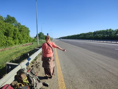 A woman stands on the shoulder of a highway in Алтынды ауылдық округі, Kazakhstan, hitchhiking. She is viewed from the back, with her right arm extended and thumb up. She wears a salmon-colored shirt, loose brown pants, and a black strap across her torso. A backpack and other luggage sit beside her near the guardrail. The highway is visible, with a row of trees on either side. Streetlights line the road. The sky is clear blue. A vehicle is visible in the distance. The scene appears to be during daylight.