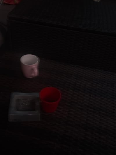 A low-angle, dimly lit photograph captures two cups and an ashtray placed on a dark, textured surface, possibly a wooden table or rattan furniture. In the upper left portion of the frame, a pink ceramic mug with a handle is partially visible, its interior appearing white. To the right of the pink mug, a red ceramic cup stands upright, also showing its interior. In the lower left quadrant, a rectangular ashtray, appears to be made of grey or silver material, is positioned on the surface. The background is dominated by a dark, woven texture, possibly a piece of furniture or wall covering. The lighting is subdued, creating a sense of shadow and depth. There are no people visible in the image. The scene is devoid of any explicit clues to its specific location within Hausach, Germany.