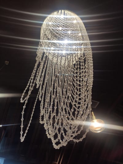 The image shows a crystal chandelier hanging from a dark ceiling. The chandelier is made of many strands of clear beads, with a larger, round glass light fixture at the bottom right. The light fixture is emitting a warm glow, and there are lens flares present due to the bright light reflecting off the crystals. The background is dark, with horizontal lines suggesting a wooden paneled ceiling or wall. The image is taken from below, looking up at the chandelier. The location is not specified in the image.