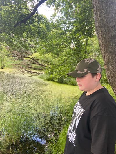 This media file depicts a young person wearing a camouflage baseball cap and a black t-shirt, standing outdoors near a body of water. The person is looking downwards and slightly to the right. The t-shirt has a white graphic design on the right side, partially visible, with bold, stylized lettering that appears to read "LIFE".
The setting is a natural environment with lush green trees overhead and to the left of the frame. The ground is covered in grass and reeds. To the right and in the background, there is a calm body of water, heavily covered with green algae or duckweed, giving it a thick, textured appearance. Some reeds and other aquatic plants are also visible along the edge and within the water. A large tree trunk is visible in the upper left corner, partially obscuring the foliage. The scene suggests a warm, possibly sunny day, with the sunlight filtering through the leaves. The overall mood is relaxed and contemplative. The provided location context is Matchino, Russia.