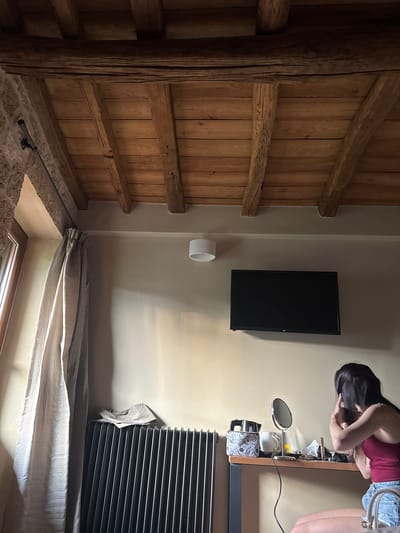 This vertical image depicts an interior room, likely within Veria Municipality, Greece, showcasing a blend of rustic and modern aesthetics.
On the left side of the frame, a dominant feature is a large wooden structure. It comprises thick vertical planks and sturdy horizontal beams, forming a wall or partition, with a substantial, textured wooden pillar or beam running vertically along the far left. The base of this wooden structure meets a rough, stone-like wall or foundation, where a dark metal pipe or rod is attached.
To the right of the wooden structure, a light-colored wall is visible. A black flat-screen television is mounted on this wall. Below the television, a white cylindrical object protrudes from the wall, possibly a light fixture or a speaker. Further to the right, a woman with long dark hair, clad in a red sleeveless top and denim shorts, is leaning forward over a wooden vanity or table. She appears to be engaged in personal grooming, possibly adjusting her hair or applying makeup, as indicated by her posture and the presence of a round, freestanding mirror and various personal items like bottles, containers, and a patterned bag on the table surface. A black electrical cord snakes across the tabletop.
Beneath the vanity, a large black radiator with horizontal fins is visible. Below the radiator and table, light-colored, folded curtains or blinds partially cover a window, letting in some ambient light. The scene is illuminated by indoor lighting, making the exact time of day indeterminate. No discernible text is visible in the image.