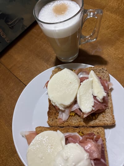 This media file depicts a close-up, overhead view of two open-faced sandwiches with prosciutto and mozzarella, served on whole wheat toast, resting on a white plate. The sandwiches are topped with slices of fresh mozzarella and appear to have a spread of mustard beneath the prosciutto. Next to the plate is a clear glass mug filled with a layered coffee drink, likely a latte or cappuccino, with a frothy top. A book or magazine with text visible lies on the wooden table in the foreground, partially obscuring the mug. The overall scene suggests a light meal or snack. The setting is a wooden surface, consistent with a table. The lighting suggests it might be daytime, though no specific time can be definitively determined. No people are visible in the image. Visible text on the book includes "POD NEZNÁMÝ..." which is likely part of a title or phrase. The location context of Červená Píska, Czechia is provided but not visually evident within the image itself.