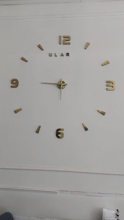 A wall clock with gold-colored numbers and a gold-colored central mechanism with a minute and hour hand is mounted on a white wall. The clock has numbers 12, 3, 6, and 9 prominently displayed, with smaller gold-colored tick marks at the intervening hour positions. The word "ULAR" is also visible, positioned below the number 12, which appears to be part of the clock's branding. The wall has decorative molding near the top and bottom. There is no indication of the city of Kalinin, Kyrgyzstan.