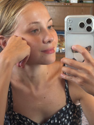 The image shows a close-up, indoor shot of a young woman taking a selfie with her smartphone. The woman has blonde hair and is wearing a black and white polka dot dress. She is holding her phone up to take the picture, and her left hand is resting on her cheek. The background appears to be a room with wooden cabinets and shelves, suggesting a domestic setting. There is no indication that this scene is located in Petryaksy, Russia.