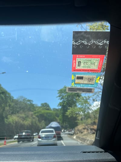 The image is taken from inside a car traveling on a road. The windshield is covered with three stickers: a black one with white text, a red one with white text, and a blue one with black text. The road is surrounded by lush green vegetation. The blue sky can be seen through the windshield. The car is traveling in a line of other cars. The image is taken from the driver's seat. The location is a road near the coordinates 9.948, -84.175 in the city of Pozos, Costa Rica.