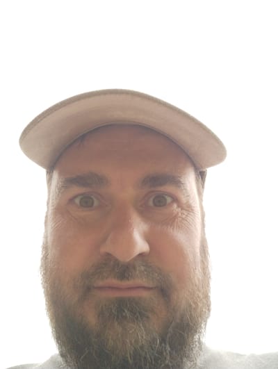 A close-up, low-angle, waist-up shot shows a man with a beard wearing a baseball cap. The man's face is centered in the frame, with his eyes looking directly at the camera. He has light brown hair and a medium-complexioned face with wrinkles around his eyes. The baseball cap is tan and tilted slightly forward. The background is a bright, blown-out white, indicating an outdoor setting with strong lighting. The man is wearing a grey shirt. The photograph appears to have been taken in the village of Shiryaevo (Il'insky), Russia.