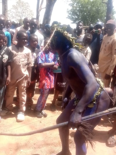 An outdoor scene in Kagoma, Nigeria, depicts a public gathering under daylight. The foreground features a male individual with dark blue body paint, wearing a leafy headdress and a sparse leafy waist covering. He is in a bent-over posture, facing towards the viewer's right, and holds a dark staff-like object with a bushy tuft near the ground. Surrounding him is a crowd of approximately twenty-five individuals, primarily men and children of various ages, observing the central figure. Most spectators wear casual attire, including shirts and trousers, with some in traditional Nigerian garments. The ground is dry, reddish-brown earth. Trees with sparse foliage are visible in the bright background. At least one male spectator in the background is holding a smartphone, potentially recording the event.
