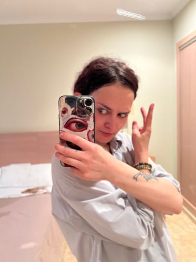 A woman is taking a selfie in what appears to be a bathroom. She has dark hair, fair skin, and is wearing a light-colored blouse. She is holding a smartphone with a white case covered in eye designs. Her left arm is visible, adorned with a bracelet and a tattoo. She is making a peace sign with her left hand. The bathroom features light-colored walls and a mirror in which the selfie is being taken. The location is Puçol, Spain, but specific details to confirm this are not visually evident in the image.