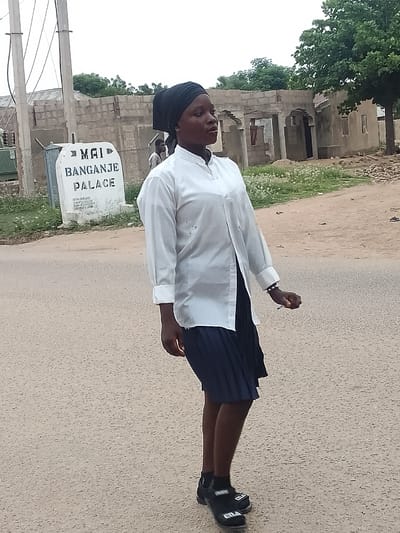 A woman in a white long-sleeved shirt and dark skirt walks on a paved road in Biliri, Nigeria. She wears a dark head covering and black shoes with visible white lettering. Behind her, a sign reads "MAI BANGANJE PALACE." The background shows a partially constructed building and a tree. Another person is visible in the background. The sky is overcast.