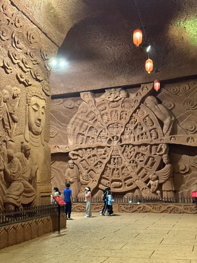The image depicts the Dazu Rock Carvings in Shizhong, Chongqing, China. The scene shows a large, intricately carved sandstone wall with Buddhist imagery, including a large reclining Buddha at the bottom and a complex mandala-like design above. Several people are standing along a barrier viewing the carvings. Three red lanterns hang near the top of the wall to the left of the mandala. The wall texture is rough and weathered, suggesting age and exposure. The environment is dimly lit, possibly artificial lighting highlighting the carvings.