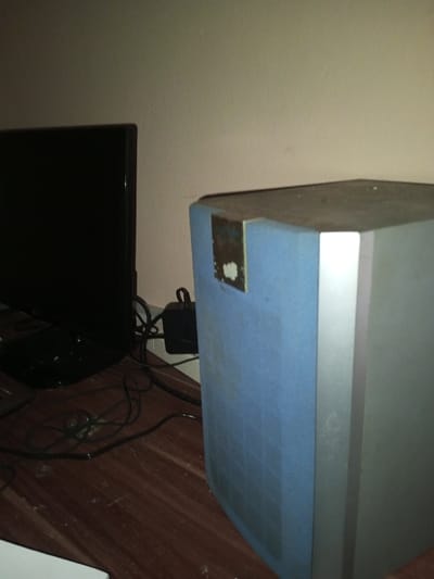 A dark screen sits on a brown surface amidst tangled black wires. To the right stands a tall, rectangular speaker with a silver side and a light blue front covered with a grid pattern. An adhesive patch, stained with remnants of a removed object, is visible on the speaker’s front. Dust covers the speaker's top and the surrounding desk surface. A black power adapter is tucked behind the speaker.