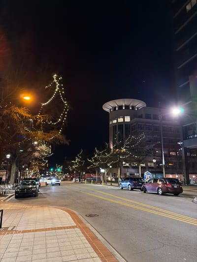 A nighttime street scene features a round building with lights in the upper left. Trees adorned with white lights line the street. Parked cars are visible on the right side of the road, adjacent to a sidewalk paved with light-colored square stones and a brick border. Streetlights illuminate the scene, casting light onto the road.