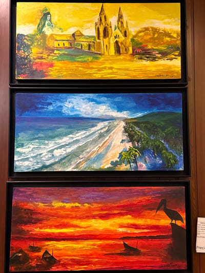 The image displays three framed paintings arranged vertically. The top painting depicts a yellowish landscape with a large, ornate building, possibly a cathedral, with spires, and a figure in green to the left. The middle painting shows a vibrant seascape with a beach, palm trees, and a dramatic blue sky with clouds. The bottom painting is a striking sunset over water, featuring silhouetted boats and a pelican perched on a rock. There is a white label in the bottom right corner with text that is partially legible, including "TELEFONO" and "Precioso." While the paintings are visually distinct, there is no information within the image that directly links them to the city of Torrejón de Ardoz, Spain.
