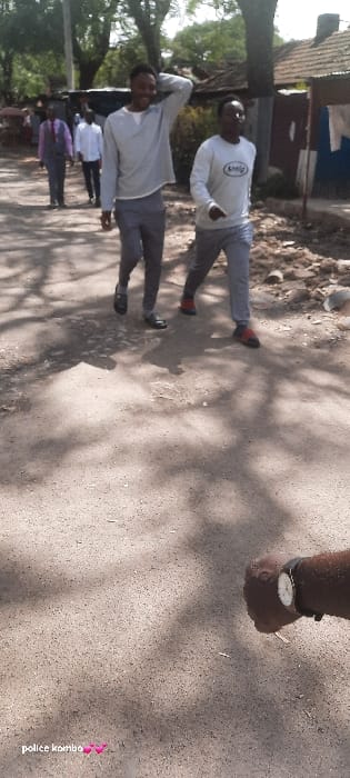 The image depicts an outdoor scene on an unpaved, dirt road in the city of Homa Bay, Kenya, under bright daylight conditions. Two men are walking forward in the mid-ground, while several other individuals are visible further back along the road.
The man on the left wears a light grey long-sleeve shirt and dark grey trousers, with black shoes. His right hand is raised to the back of his head/hair, and he is smiling, looking towards the camera's left. The man to his right wears a white long-sleeve shirt with a dark oval-shaped logo on the chest, dark grey trousers, and dark shoes with visible red accents (possibly socks). He walks with his arms at his sides, looking forward.
In the background, behind these two individuals, additional people are walking away from the camera. One person wears a grey suit jacket over a purple shirt, while another wears a white shirt.
The road surface is light brown, dusty soil with visible small stones and scattered dark shadows, likely from overhead trees. To the right of the road, the ground appears uneven and littered with debris. Buildings with various roof types, including corrugated iron and reddish-brown tiles, are partially visible along the road, suggesting a semi-urban or residential area. Green foliage from trees is visible in the upper background.
In the bottom right corner of the frame, a dark-skinned forearm and hand, wearing a watch with a white face and black strap, is partially visible, indicating the presence of another person or the photographer at the immediate foreground.