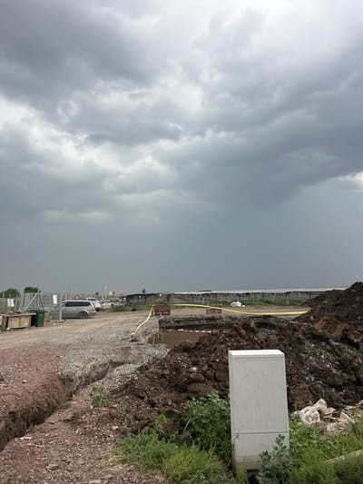 An overcast day in Tenevo, Bulgaria, looms over a construction site. The sky is filled with heavy, gray clouds, suggesting impending rain. In the foreground, a trench has been dug into the earth, with piles of dark soil and gravel surrounding it. A white electrical box stands near the edge of the construction zone, nestled among green vegetation. Beyond the immediate construction area, a dirt road leads past several parked vehicles, including a silver SUV and a white car. In the distance, a long row of solar panels stretches across the landscape, a testament to the ongoing development in the region. The overall scene depicts a site undergoing active construction and development under a somber sky.