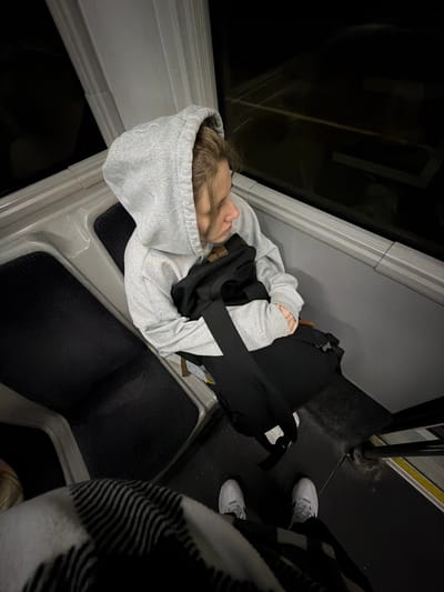 The scene depicts the interior of a transport vehicle, likely a bus or train, in dim lighting conditions. A single individual, appearing to be a young adult with light-colored hair, is seated on the right side of the frame. This person wears a light grey hooded sweatshirt with the hood up and dark trousers, and is looking towards the left, slightly downward. A black bag with a visible strap is positioned across their lap. To their left, an identical, unoccupied dark-textured seat with a light grey frame is visible. The vehicle's interior features light grey paneling along the wall, below a dark window that reflects minimal external light, suggesting a dark outdoor environment. The floor is dark. In the immediate foreground, the lower legs and white sneakers of a second individual are visible, standing on the dark floor. This second individual is partially covered by a white and black striped or checkered blanket or garment. No visual identifiers within the image provide information regarding a specific geographical location, such as the city of Boldino, Russia.