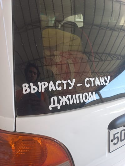 The image shows the rear of a white vehicle, likely in the city of Yorkatay, Uzbekistan, based on the context provided. A white sticker with Russian text is applied to the rear window. The text reads "ВЫРАСТУ - СТАНУ ДЖИПОМ" which translates to "I will grow up - I will become a jeep." The reflection in the window shows a man's face, possibly the owner of the vehicle, and a light-colored object, possibly a blanket or fabric, inside the car. To the right, a license plate with the characters "50" is partially visible. The scene appears to be outdoors, possibly in a parking area or on a street.