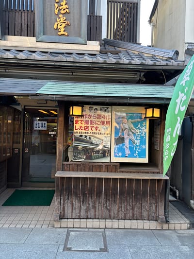 The image shows the exterior of a small shop located on a narrow street in Katsushika City, Tokyo, Japan (latitude: 35.758, longitude: 139.877). The shop's facade is made of wood and features a green awning with Japanese characters. The awning is attached to a narrow, wooden structure that serves as a covered entrance to the shop. The entrance is framed by two brown, wooden pillars, and a wooden platform that is covered in black and white tiles. The platform sits just above the level of the street, which is paved with gray and beige square tiles. There is a green mat on the left side of the entrance and a small, square, gray plate on the right side. The shop’s windows are covered with posters, including one featuring a man in a blue jumpsuit standing with his arm raised in the air. Other posters feature text written in Japanese. The shop’s window frames are made of wood. There are two large, white fluorescent lights that illuminate the shop's interior. The shop appears to be closed, and there are no people present.