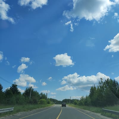 The image shows a view from a vehicle traveling on a paved road with a yellow lane divider. The road is surrounded by green grass and trees on both sides. In the distance, there is a dark-colored SUV driving ahead. Power lines and utility poles are visible along the roadside. The sky is blue with scattered white clouds. There are no people visible in the image. The scene appears to be a rural or semi-rural area. The location in Kineshma, Russia, cannot be determined from the visual information provided in the image.