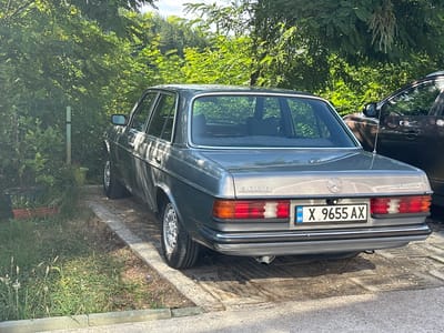 The image shows the rear of a silver Mercedes-Benz W123 sedan parked on a concrete surface surrounded by grass and trees. The car has Bulgarian license plates reading "X 9655 AX" and the emblem "300D" and "AUTOMATIC". A dark colored SUV is parked to the right of the Mercedes. The environment appears to be outdoors, possibly in a residential area or a parking lot, given the presence of a fence post and plants. The lighting suggests it is daytime, with sunlight filtering through the leaves of the trees. The car is clean and appears to be in good condition. There are no people visible in the image.