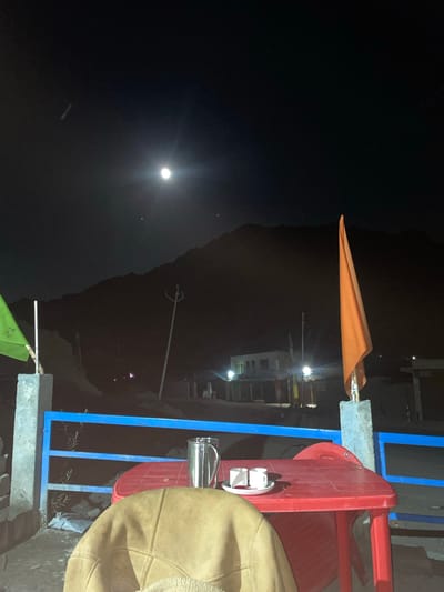 This is a night-time photograph taken outdoors, likely in the city of Khangkor, India, featuring a view of a moonlit mountain range. In the foreground, a red plastic table is set with a metal pitcher and two small white cups on a white saucer, suggesting a beverage service. A beige, possibly leather or suede, jacket or coat is draped over a chair in the lower part of the frame. A blue metal railing separates the foreground area from a lower level, where some debris and vegetation are visible. Two flags are present: a green flag on the left, partially obscured, and a prominent orange flag on the right. Behind these elements, a dark mountain silhouette is illuminated by the full moon, which is visible in the dark sky. Some distant lights suggest the presence of buildings and infrastructure in the mid-ground, possibly a small settlement or roadside establishment. The overall scene conveys a quiet, possibly cold, outdoor evening setting.