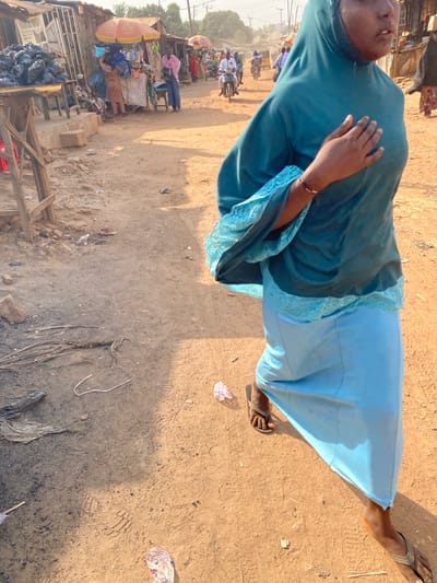 A woman in a blue dress and sandals stands on a sandy, littered ground. A market scene unfolds in the background with people, motorbikes, and stalls under umbrellas. Trash bags and remnants of burnt materials are scattered nearby. Sunlight casts strong shadows on the ground, highlighting the details of the rough terrain. The woman is seen from a slightly elevated perspective.