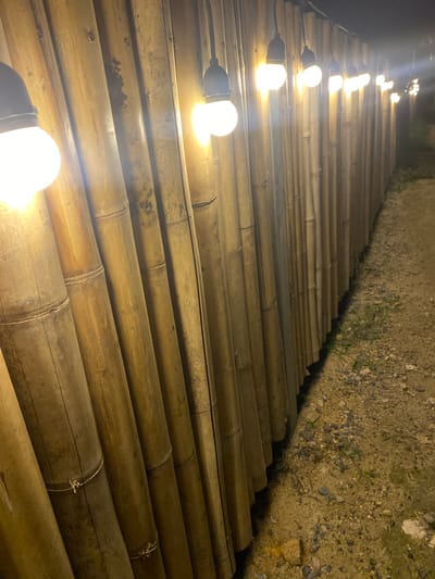 The image depicts a nighttime scene in Baan Don Sai, Thailand. A pathway is constructed of numerous bamboo poles laid side-by-side, creating a walkway. A string of lights illuminates the path, with each bulb emitting a warm, bright glow. The ground on one side of the pathway is dirt and gravel, and the opposite side fades into darkness. No people are visible. The focus is on the illuminated path and the construction materials used.
