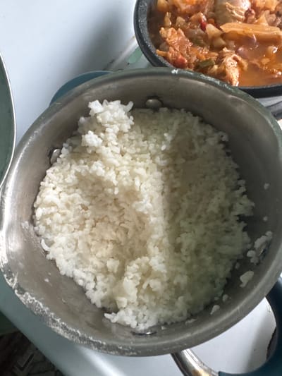 A close-up, overhead shot captures a pot filled with fluffy white rice. The pot is silver-toned and shows signs of use. To the right, a portion of another pot is visible, containing a hearty stew or curry with visible pieces of meat and vegetables in a reddish-orange sauce. The background appears to be a kitchen counter. The image does not contain information to determine the location within the city of Krasnoe, Russia.