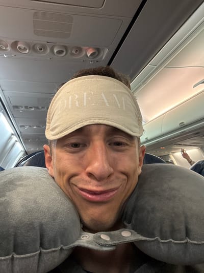 A man wearing a "DREAM" sleep mask and a grey travel pillow poses for a selfie on an airplane. The plane's interior, including overhead vents and passenger seats, is visible. The lighting suggests daytime. The man's expression is a slight smile, and his head is tilted to one side. His neck pillow is secured with button closures.
