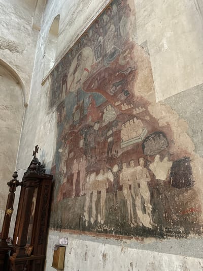 The image shows a large, well-preserved fresco on the wall of a stone building. The fresco depicts a religious scene with multiple figures, some with halos, and is executed in a traditional, somewhat stylized manner with earthy colors. At the top of the fresco, a central figure, likely Christ, is depicted within a circular halo, surrounded by other figures. The rest of the fresco appears to illustrate various narratives, with groups of people, animals, and possibly scenes of judgment or historical events. The wall itself is made of rough-hewn stone blocks, and there are architectural elements such as arched windows visible in the background. To the left of the fresco, a dark wooden structure with intricate carvings is present, topped with a cross, suggesting it is part of the church's interior furnishings. The overall environment appears to be an ancient religious site, with the fresco serving as the primary focal point. The location is identified as Shalikiantkari, Georgia.