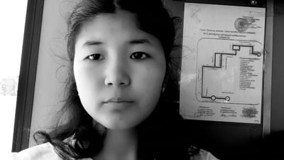 A black and white, medium close-up shot shows a young East Asian woman's face from the nose down, looking directly at the camera. Her dark hair is loose around her face and shoulders. Her eyes are dark and wide, and she has a neutral expression. A small earring is visible on her right ear.
To the right of the woman is a partially visible poster or map, which appears to be a bus route diagram. The text on the poster is in Cyrillic script. The top section of the poster has text that reads "Жаны-Кыштак, Кыргызстан" and "автобустун схемасы" (bus route map). Below this is a diagram of bus stops and routes. The bottom of the poster is obscured by the woman's shoulder and hair.
The background is a dimly lit, indoor environment, possibly a bus interior, with a hint of what might be a window or door frame on the left side.