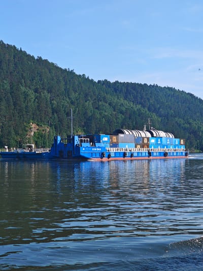 The image shows a large blue barge carrying numerous shipping containers and large cylindrical objects on a wide river. The barge is moving from left to right. In the background, a dense green forest covers a steep hillside, and a clear blue sky is visible above. The water in the foreground is dark and reflects the trees and sky, with some small waves and foam visible on the right side. A smaller, lighter-colored barge is attached to the left of the main barge. The main barge has Cyrillic lettering on its side that reads "БЛМ-2503". The scene appears to be of the Yenisei River near Predivinsk, Russia, a town known for its river port and transportation of goods along the waterway.