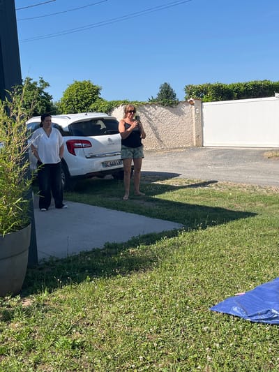 The image captures an outdoor scene in Saint-Just-Saint-Rambert, France, under a clear blue sky with strong sunlight casting distinct shadows. Two adult females are present. On the left, a woman with dark hair tied back, wearing a white V-neck top and dark pants, stands on a gray concrete path with her hands in her pockets. To her right and slightly behind her, a second woman, wearing sunglasses, a dark tank top, and patterned shorts, stands on a gravel-like surface. This woman is holding a black smartphone horizontally with both hands, pointed towards the left.

Behind the two women is a white Citroën C3 Aircross SUV, parked facing away from the viewer, displaying a French license plate (DC-440-LR). The background features a textured light-colored stone wall with a tiled coping, behind which various green trees and bushes are visible. A white metal gate is positioned on the right side of the background wall. The foreground consists of green grass interspersed with small white flowers, partially shaded by an unseen object to the left. A large potted plant with tall green stalks is visible in the lower-left corner. A blue tarp or folded fabric is located on the grass in the lower-right foreground. Faint power lines traverse the upper portion of the clear blue sky.