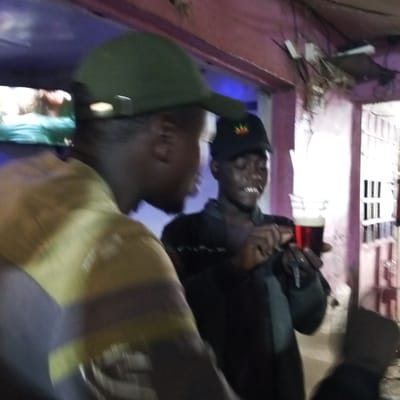 The image depicts two men in what appears to be a bar or informal gathering place in Kinoo ward, Kenya. One man, partially obscured, is wearing a green cap and a yellow and brown striped shirt. Another man is wearing a black cap with a red and yellow emblem. He is holding a glass containing a dark red liquid, possibly beer or soda, and is looking at it with a slight smile. The background shows a partially visible doorway with vertical metal bars, purple painted walls, exposed electrical wires, and other individuals, partially visible. The scene is dimly lit.