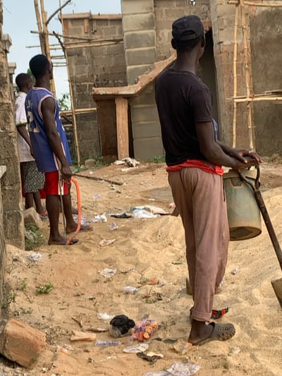 A man in a black shirt, brown pants, and a cap is carrying a container using a metal bar. Behind him, a building under construction is visible. In the foreground, two men are working with a long, orange saw-like tool. The ground is covered in sand and strewn with various pieces of debris, including paper and plastic bags. One man wears a blue jersey, red shorts, and sandals. Another man is standing beside him with an unseen attire.