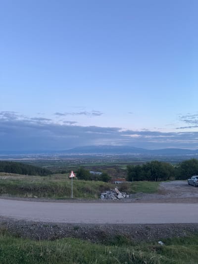 The image captures a panoramic landscape view of a valley and distant mountains in Balsha, Bulgaria, during twilight, suggesting either early morning or late evening. The sky, occupying the upper portion, is predominantly clear and blue, transitioning to a lighter hue towards the horizon, with scattered, elongated clouds.
In the middle ground, an expansive green valley is visible, dotted with what appear to be distant settlements or a small city, marked by faint patterns of buildings. Beyond the valley, a chain of mountains rises, appearing hazy and soft against the horizon.
The foreground on the right side of the image features a partially unpaved or gravel road running through a grassy area. To the left of the road, a triangular warning sign with a red border, white interior, and a black exclamation mark stands on a pole. The terrain immediately surrounding the road consists of green grass, gravel, and scattered rocks. On the far right, near the top, a portion of a white vehicle, possibly a van or camper, is visible, parked near some trees. No specific activity or event is taking place; it is a static landscape scene. No discernible text is visible other than the exclamation mark symbol on the warning sign.