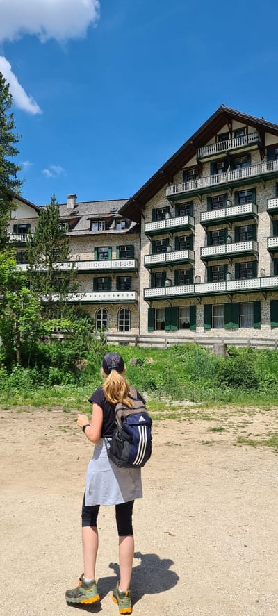 A woman with blonde hair pulled back in a ponytail, wearing a black t-shirt, grey skirt, black leggings, a black cap, and hiking boots, stands facing away from the camera. She is carrying a blue and grey backpack with white stripes. She appears to be walking on a dirt path in front of a large, stone building with multiple balconies and green shutters. The building is surrounded by trees and lush green grass. The sky is blue with scattered white clouds. The scene suggests a location in the city of Prags - Braies, Italy, likely a hotel or resort area in a mountainous region.