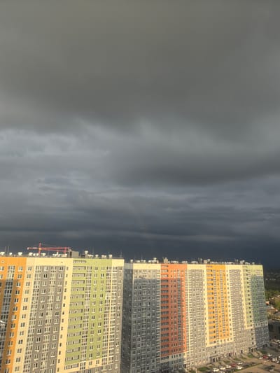 A ground-level, outdoor, medium shot, depicts a dark, stormy sky next to a cityscape with multiple apartment buildings, in Ankudinovo, Russia. The sky is predominantly gray and overcast, suggesting an approaching storm. The apartment buildings are tall, with a grid-like pattern of windows, colored with gray, yellow, orange, and green. Cars are parked along the street next to the buildings. A construction crane is visible near one of the buildings.