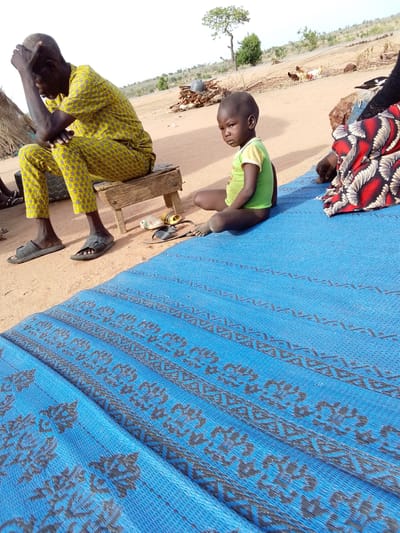 The image depicts an outdoor scene in Mulke Barikin Sayejo, Nigeria, with several people present. A man in a yellow patterned outfit sits on a small wooden stool, his head in his hand. A young child in a green shirt sits on a blue patterned mat. A person wearing a red and white patterned garment sits nearby. A pile of wood and several chickens are visible in the background. The ground is sandy and dry.