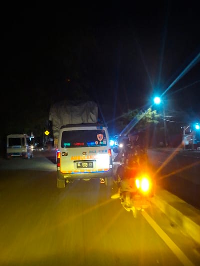The image depicts a nighttime scene on a road, likely in Lamprehlam Teungoh, Indonesia. The primary focus is an ambulance with the license plate "BL 8138 ZJ". The ambulance is white with red and blue emergency lights visible on its rear. To the left of the ambulance, another vehicle, possibly a van, is visible, also illuminated by headlights. Further down the road, another vehicle with flashing lights is seen. A scooter with a bright headlight is in the foreground, obscuring much of the road surface and creating a significant glare. The environment appears to be a relatively dark road with some trees and streetlights visible in the background. The overall impression is of a convoy or a traffic scene at night.