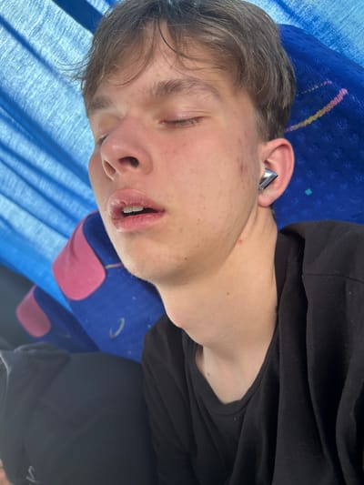 A young man is asleep on what appears to be a bus seat. He is lying on his side, with his head resting on the blue and pink patterned headrest. He has fair skin, light brown hair, and is wearing a black hoodie. His mouth is slightly open, and an earring is visible in his left ear. The location is unknown, and the city of Krushare, Bulgaria, is not evident in the image.