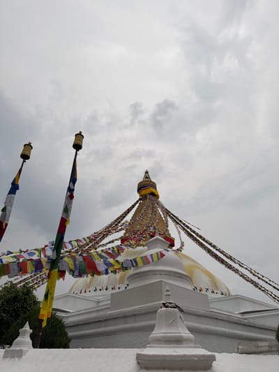 A low-angle shot of the Boudhanath Stupa, located in the city of Gokarneshwar, Nepal, taken on an overcast day. The stupa's white dome is partially visible on the right side of the frame, with its distinctive square tower above. Colorful prayer flags are draped from the tower, extending outwards. Three ornate metal cylinders, adorned with prayer flags, are positioned diagonally in the foreground. The backdrop is a gray, cloud-filled sky.