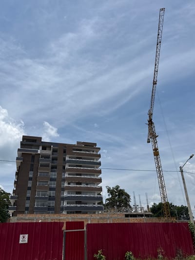 The image displays a construction site in Pitalito, Colombia, under a partly cloudy sky. A multi-story building, estimated to be ten to twelve floors, is under construction, featuring alternating sections of dark brown and gray cladding, with balconies on various levels that have white railings. The upper floors appear less complete, showing exposed structural elements.

To the right of the building, a tall, yellow lattice-boom tower crane stands prominently, its mast extending significantly above the building's current height. In the foreground, a red corrugated metal fence, approximately two meters high, encloses the construction area. A smaller, hinged gate section is visible on the left side of this fence. A circular white sign with a red border and a black 'P' (likely indicating parking regulations) is affixed to the left portion of the fence. Behind the fence and in front of the main building, visible lower concrete and brick structures with exposed rebar indicate ongoing foundational or ground-level construction.

On the far right, a utility pole with an attached street lamp and overhead wires is visible. Green tree foliage is present in the background and midground. No individuals, specific actions, or interactions are discernible within the frame.