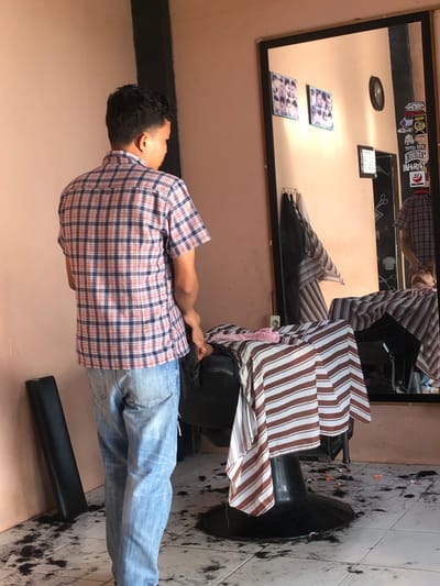 A person wearing a plaid shirt and blue jeans sits in a barber chair covered with a striped cape. The barber shop is in Pulorayeuk, Indonesia. Hair clippings are scattered on the tiled floor. A mirror hangs on the wall, reflecting the person in the barber chair, as well as some items on the wall. These include stickers, pictures and a clock. A black footrest is visible near the chair.