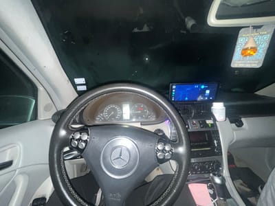 The image provides a detailed view of the interior of a Mercedes-Benz car, likely a C-Class (W203 generation), at night. The perspective is from the passenger side, looking towards the driver's area and the dashboard.

The steering wheel, centered in the frame, is black with visible stitching and features a Mercedes-Benz logo in the center. It has multi-function controls on both sides, with four circular buttons on each side. "SRS AIRBAG" is embossed on the lower part of the steering wheel hub. Behind the steering wheel, the instrument cluster displays a speedometer on the left (up to 260 km/h) and a tachometer on the right. A digital display is visible between the gauges, and an illuminated engine warning light is present on the right gauge.

Mounted on the dashboard, above the center console, is a large electronic display showing a map application with the time "22:30" and icons for "Search" and "ATM." The map displays road networks and includes faint text such as "Kresna," a town in Bulgaria. Below this display, the car's original infotainment unit and climate controls (featuring three rotary knobs) are visible. A manual gear shifter with a silver trim is located to the right of the steering wheel, closer to the passenger side. A pink sticky note with handwritten text "10.06" and "12:30" is placed near the gear shifter.

On the dashboard, near the passenger side, a white plastic bottle (possibly supplements or medication) is placed. An air freshener, containing orange liquid in a wooden top, hangs from the rearview mirror. Its label features a QR code, Bulgarian text "Свали си ТУК" (Download HERE), the website "www.krisauto.bg", and a Bulgarian phone number "0887 400 004". The car's interior pillars are light grey. A small spider is visible on the passenger-side A-pillar, above the steering wheel. The exterior is dark, indicating nighttime. A portion of a leg, wearing grey pants, is visible on the far right of the frame, suggesting a passenger is present.

While the presence of Bulgarian text and a Bulgarian phone number on the air freshener indicates a location within Bulgaria, the specific city of Elhovets cannot be definitively confirmed from any visible details within the image, as the map application shows other Bulgarian towns like Kresna.