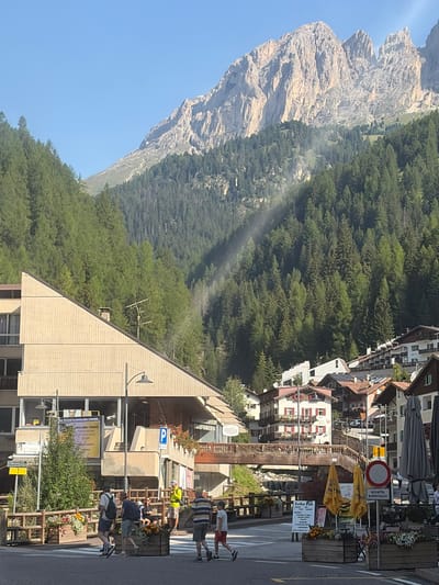 The image depicts a sunlit street scene in the Italian town of Campitello di Fassa, nestled in the Dolomites. The backdrop is dominated by the imposing, rocky peaks of the mountains, partially obscured by a dense evergreen forest that covers the slopes. A faint, misty beam of sunlight cuts through the trees, creating an ethereal effect.
In the foreground, a street in Campitello di Fassa is visible. A modern, angular building with a light-colored facade stands to the left, featuring multiple levels and a large, angled roof. Adjacent to it, a sign reads "PARUCCHIERE PROFUMERIA," indicating a hair salon and perfumery. A wooden pedestrian bridge crosses a small stream or ravine in the mid-ground, connecting different parts of the town.
Several people are present in the scene. Two men in casual attire are walking across a crosswalk on the left side of the street, one with a backpack. Another man in a bright yellow jacket stands further down the street, observing. A couple is seated on a bench to the right, near a planter box filled with colorful flowers. Further to the right, two children are walking, one in a red shirt and shorts, and another in a striped t-shirt and shorts.
Various objects are scattered throughout the scene, including streetlights, planter boxes with flowers, yellow umbrellas shading outdoor seating areas, and traffic signs, including a "no entry" sign and a sign with Italian text. The architecture of the town features traditional alpine style, with tiled roofs and wooden balconies. The overall atmosphere is one of a peaceful, sunny day in a charming mountain town.
