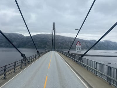 This is a forward-facing, wide-angle shot of a suspension bridge in Sandnessjøen, Norway. The bridge's roadway, marked with yellow lines, stretches into the distance, curving slightly as it ascends towards the central tower. The tower itself is a tall, slender structure with cables fanning out to support the bridge deck.
On the right side of the road, a lamppost stands near a triangular warning sign. The sign depicts a car skidding on a wet surface and below it reads "Ved våt vegbane" and "40 KM/T", indicating a speed limit of 40 kilometers per hour for wet roads. A metal railing lines the side of the bridge, separating the roadway from the dark, still water below.
In the background, imposing, rocky mountains rise, their peaks shrouded in low-lying clouds. The weather appears overcast and potentially damp, contributing to the subdued atmosphere. The overall scene conveys a sense of vastness and isolation, with the bridge serving as a man-made artery connecting landmasses amidst a rugged, natural landscape. There are no people visible in the image. The time of day is indeterminate due to the cloudy conditions.